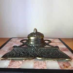Könnte beinhalten: Ein antikes Tintenfass aus Messing mit einem zentralen Reservoir mit Deckel, das mit komplizierten floralen und geometrischen Reliefmustern versehen ist. Die Basis enthält zwei breite, flache Stiftablagen mit detaillierten Verzierungen.