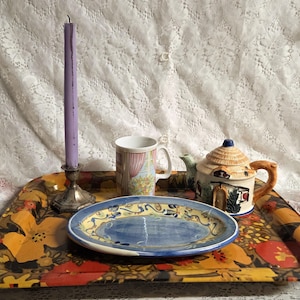Puede incluir: Una bandeja decorativa con un diseño floral sostiene una vela morada, una taza, un plato azul y blanco y una tetera. La tetera tiene un diseño similar a una casa. La bandeja es rectangular con un colorido estampado floral.