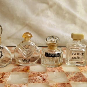 Pode incluir: Uma coleção de cinco frascos de perfume vintage de várias formas e tamanhos. Os frascos são de vidro transparente com detalhes e rótulos dourados. Os frascos estão dispostos sobre uma superfície decorativa quadriculada.
