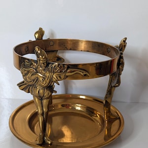 May include: A vintage brass wine bottle holder with a circular design. The holder features three decorative figures in period clothing, supporting a ring that holds the bottle. The base is a round, shallow dish. The overall color is gold.