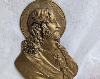 May include: A bronze-colored religious relief sculpture depicting a figure with long hair, a beard, and a halo. The figure is wearing draped clothing and has a heart symbol on the chest. The sculpture is likely made of metal.