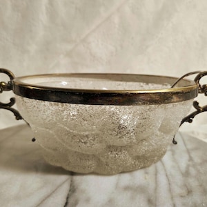 Puede incluir: Un cuenco de vidrio transparente con un aspecto texturizado y esmerilado. El cuenco tiene un borde dorado y asas doradas ornamentadas. El cuenco es probablemente vintage y adecuado para servir.