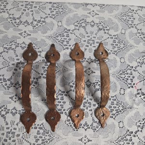 May include: Four copper-toned door handles with a hammered finish. Each handle features a decorative top and bottom with a mounting hole. The handles are displayed vertically against a white lace backdrop.