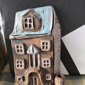 Puede incluir: Figurita de casa de cerámica hecha a mano, con tejado azul claro y paredes marrones. La casa tiene varias ventanas y una pequeña chimenea. Un pequeño objeto en forma de barril está al lado de la casa. La figurita está sobre una superficie a rayas verdes y blancas.