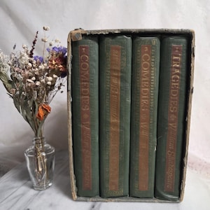 May include: A collection of four antique green books in a cardboard box. The spines display gold lettering: "Comedies & William Shakespeare" and "Tragedies & William Shakespeare". A small glass vase with dried flowers is positioned to the left.