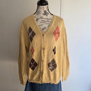 May include: Mustard yellow argyle cardigan sweater with a V-neck and button-up front. The sweater features diamond patterns in brown, white, and coral. The sweater is displayed on a mannequin with a denim skirt.