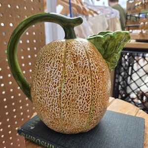 May include: A ceramic jug shaped like a cantaloupe melon with a textured, tan and brown netted rind. The handle is a curved green vine, and a green leaf extends from the top near the spout. The jug has a rounded, bulbous body.
