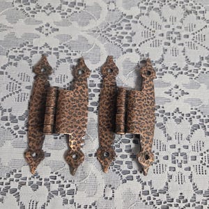 May include: Two antique copper-colored hinges with a hammered texture. Each hinge has a decorative shape with pointed ends and a series of barrel-shaped sections. The hinges are designed for a door or cabinet.