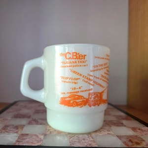 Op de afbeelding: Een witte melkglazen mok met oranje tekst en illustraties gerelateerd aan CB-radioterminologie. De opdruk bevat termen zoals the C.B'er, TIJUANA TAXI, GREEN STAMP HIGHWAY, FLIP FLOP en 10-4, vergezeld van grafische afbeeldingen van een auto en een vrachtwagen.