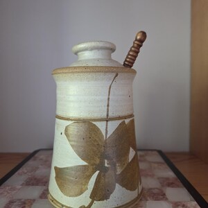 Peut inclure: Un pot à miel cylindrique en céramique avec un vernis blanc cassé moucheté et un motif floral marron peint sur le devant. Le pot comprend un couvercle assorti avec une petite ouverture et une cuillère à miel en bois insérée dans le haut.