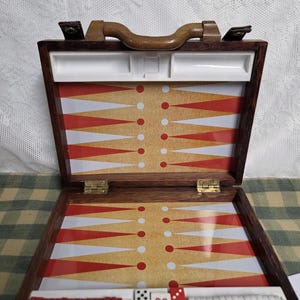 Puede incluir: Juego de backgammon de madera abierto con superficie de juego roja, blanca y beige. El juego incluye fichas rojas y blancas, dados y un cubo de duplicación. El estuche tiene un asa de madera y bisagras de latón.