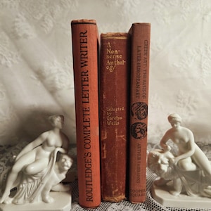 Könnte beinhalten: Drei antike Bücher, darunter "Routledge's Complete Letter Writer" und "Greek Art: The Basis of Later European Art", stehen zwischen zwei weißen Keramikskulpturen einer Frau, die einen Löwen reitet.