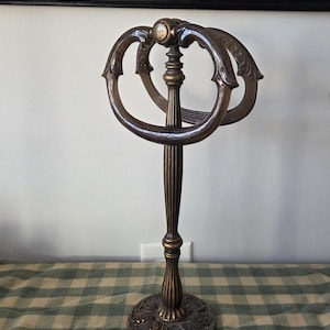 May include: An antique bronze towel stand with a decorative base and a central column. The top has two curved, ornate rings. The stand is designed to hold towels and adds a touch of vintage elegance to any bathroom.