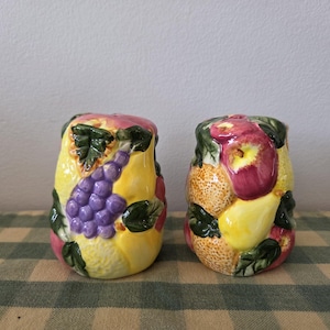 May include: A pair of ceramic salt and pepper shakers. One shaker features a cluster of purple grapes and yellow pears. The other shaker has a design of red apples and pears. Both shakers have green leaves.