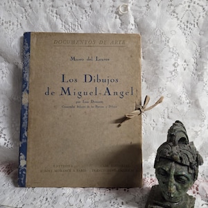 Puede incluir: Un libro antiguo titulado "Los Dibujos de Miguel-Angel" con texto azul y una cubierta marrón, junto a una pequeña escultura verde de una cabeza. El libro es del Museo del Louvre y tiene una cinta de papel atada.