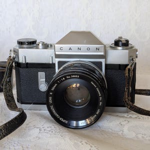 May include: A classic Canon camera, black and silver, with a 50mm lens. The lens is marked "SUPER-CANOMATIC LENS R 50mm 1:1.8". It features a leather strap and a small black case. The name "CANON" is printed on the top of the camera.