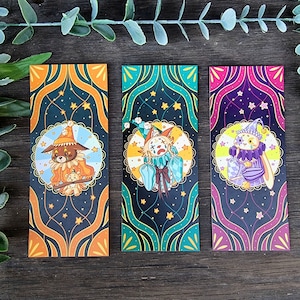 Peut inclure: Trois marque-pages illustrant des animaux habillés en clowns. Chaque marque-page a un schéma de couleurs différent et un design animal unique. Le premier marque-page représente un chien brun et blanc, le second un chat blanc et bleu, et le troisième un lapin blanc.