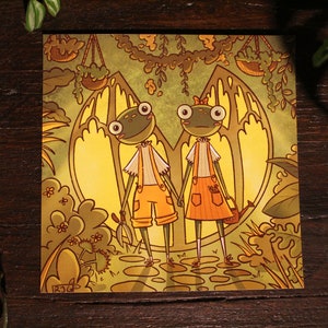 May include: Two green frog characters with large eyes and orange clothing stand in a yellow and green forest setting. The frog on the left holds gardening shears and the frog on the right holds a watering can. The background features a large yellow archway with hanging plants.