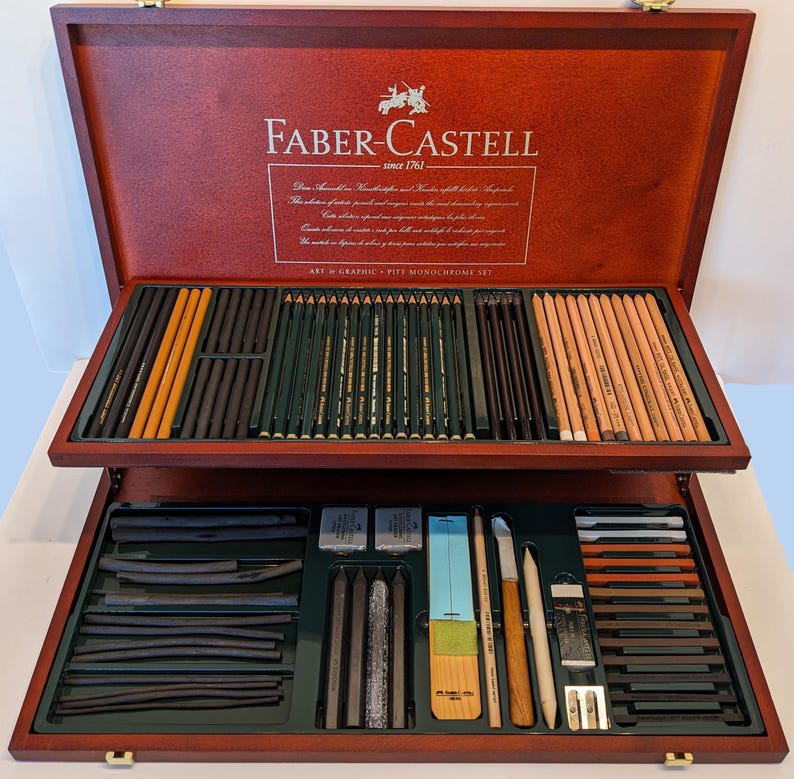 May include: A Faber-Castell Art & Graphic Pitt Monochrome Set in a wooden case. The set includes pencils, charcoal sticks, erasers, and blending tools. The case is a dark reddish-brown color with the Faber-Castell logo and text on the lid.