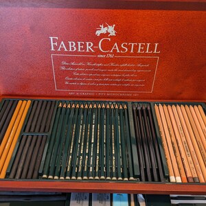 May include: A wooden Faber-Castell art set, open to reveal an assortment of pencils and charcoal sticks. The set includes various shades of pencils, including black, brown, and natural wood colors. The box is a rich, reddish-brown color, with the Faber-Castell logo and text.