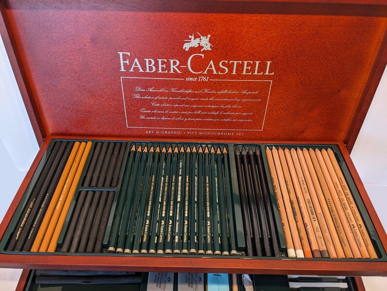 May include: A wooden Faber-Castell art set, open to reveal an assortment of pencils and charcoal sticks. The set includes various shades of pencils, including black, brown, and natural wood colors. The box is a rich, reddish-brown color, with the Faber-Castell logo and text.