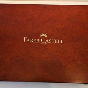 May include: A rectangular, wooden box with a rich, reddish-brown finish. The box features the gold-colored text "FABER-CASTELL" with a crest and the text "since 1761" in a gold color. The box has gold-colored hinges.