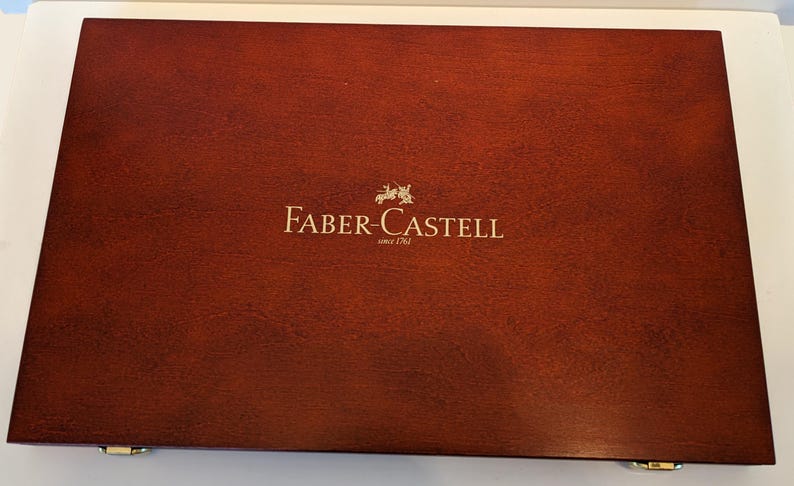 May include: A rectangular, wooden box with a rich, reddish-brown finish. The box features the gold-colored text "FABER-CASTELL" with a crest and the text "since 1761" in a gold color. The box has gold-colored hinges.