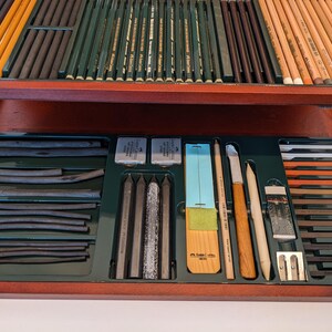 May include: A wooden art supply box with various drawing tools. The box contains charcoal sticks, pencils, erasers, blending stumps, and sharpeners. The pencils are in shades of brown, green, and yellow. The box is open, revealing the organized contents.