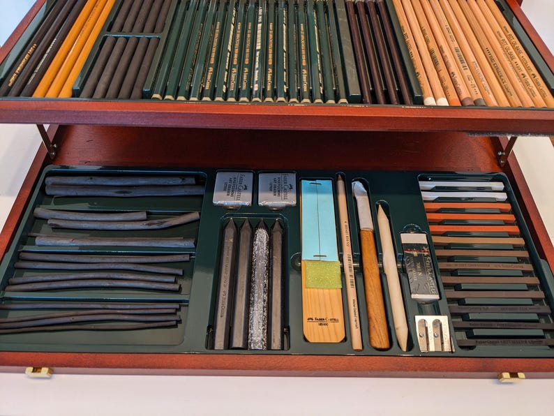 May include: A wooden art supply box with various drawing tools. The box contains charcoal sticks, pencils, erasers, blending stumps, and sharpeners. The pencils are in shades of brown, green, and yellow. The box is open, revealing the organized contents.
