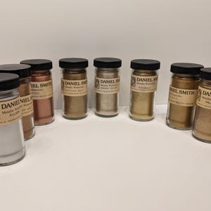 May include: A collection of glass jars filled with metallic watercolor pigments. The jars have black lids and labels with the brand name "Daniel Smith." Colors include silver, bronze, gold, and copper tones. The jars are arranged on a white surface.