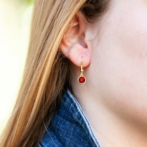 May include: A pair of gold earrings with a small red gemstone dangling from each earring.