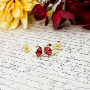May include: A pair of gold stud earrings with red gemstone accents. The earrings are on a piece of paper with handwritten text.
