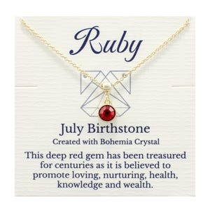 May include: A gold-tone chain necklace featuring a round, deep red crystal pendant. The display card reads: Ruby, July Birthstone, Created with Bohemia Crystal, This deep red gem has been treasured for centuries as it is believed to promote loving, nurturing, health, knowledge and wealth.