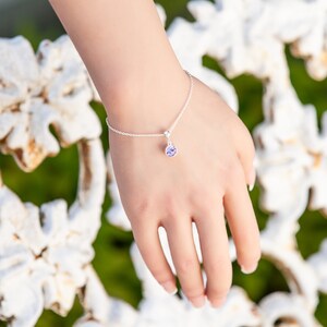 May include: A delicate silver bracelet with a small, round, light purple charm. The bracelet is on a person's wrist, with the hand in focus. The background is blurred, with white and green tones.