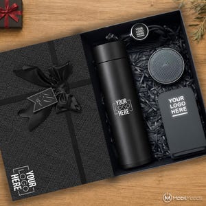 May include: A black gift box containing a matte black vacuum insulated bottle, a circular wireless speaker, and a rectangular power bank, all featuring the text YOUR LOGO HERE. The box includes a black ribbon, a gift tag, and black paper filler.