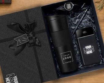 May include: A black gift box containing a black insulated tumbler, a black power bank with a digital display showing 100%, a black pen, and a black circular keychain, all featuring the text YOUR LOGO HERE, with a black ribbon and gift tag.