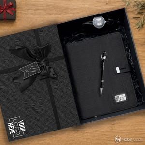 May include: A black gift box containing a textured black notebook with a black pen, a circular logo tag, and a black ribbon bow with a tag reading Just For You. The box lid features a white logo reading YOUR LOGO HERE and a black ribbon.