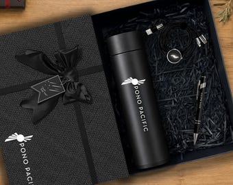 Könnte beinhalten: Eine schwarze Geschenkbox mit einer schwarzen Isolierflasche, einem schwarzen Kugelschreiber und einem schwarzen Ladekabel, die alle das Pono Pacific-Logo tragen. Der Deckel ist mit einer schwarzen Schleife und einem Just For You-Anhänger versehen.