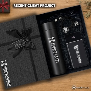 Op de afbeelding: Een zwarte geschenkdoos met een matzwarte vacuümgeïsoleerde fles, een rechthoekige powerbank met een digitaal display dat 100% aangeeft en een zwarte oplaadkabel. Elk item is voorzien van het witte Eastchurch Construction logo.
