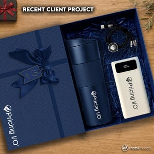 May include: A navy blue gift box containing a navy blue insulated tumbler, a white power bank with a digital display showing 100%, and a black charging cable. The tumbler and power bank feature the Pricing I/O logo. A navy ribbon is on the box.