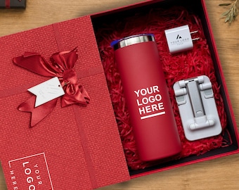 Könnte beinhalten: Eine rote Geschenkbox mit einem roten Becher mit dem Text YOUR LOGO HERE, einem weißen Netzteil mit dem Text YOUR LOGO HERE und einem weißen faltbaren Handyhalter. Der Deckel ist mit einer roten Schleife und einem weißen Etikett versehen.