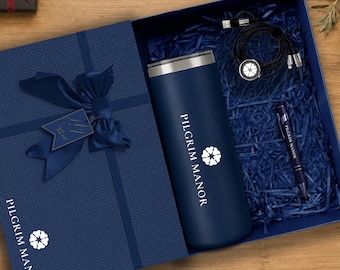 Pode incluir: Uma caixa de presente azul-marinho contendo um copo azul-marinho correspondente com o texto PILGRIM MANOR e um logotipo de flor, um cabo de carregamento preto com um conector circular iluminado e uma caneta azul-marinho com o texto PILGRIM MANOR.
