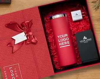 Könnte beinhalten: Eine rote Geschenkbox mit einer großen Schleife und einem Anhänger, die einen roten Becher mit dem Text YOUR LOGO HERE, einen weißen Netzadapter mit Logo und ein schwarzes, rechteckiges kabelloses Ladepad mit Logo auf rotem Füllmaterial enthält.