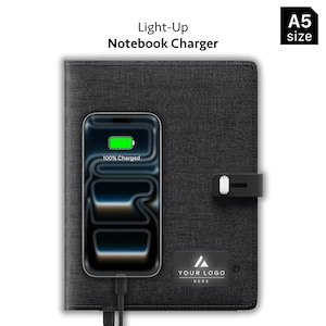 May include: A black A5 size Light-Up Notebook Charger with a textured fabric cover. A smartphone is placed on the front, displaying a green battery icon and the text 100% Charged. A black rectangular patch features the text YOUR LOGO HERE.