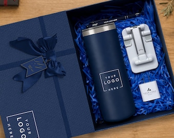 Pode incluir: Uma caixa de presente azul-marinho com fita e etiqueta a condizer, contendo um copo azul-marinho com o texto YOUR LOGO HERE, uma caneta azul-escura, um suporte dobrável branco para telemóvel e um adaptador de corrente branco com o texto YOUR LOGO HERE.