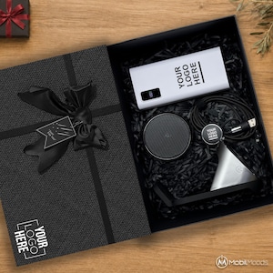 May include: A black gift box containing a white power bank with the text YOUR LOGO HERE, a circular black speaker, a black charging cable, and a silver phone stand. The box lid features a black ribbon, a tag reading Just For You, and a logo.
