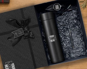 Pode incluir: Uma caixa de presente preta contendo um copo térmico preto fosco com o texto YOUR LOGO HERE, uma caneta esferográfica preta com o texto YOUR LOGO HERE e um acessório tecnológico circular preto com cabo, tudo sobre uma base de papel picado preto.