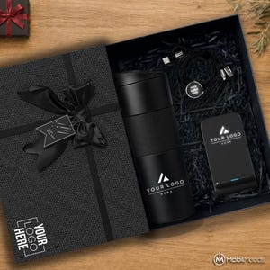 May include: A black gift box containing a black travel mug, a black wireless charging stand, and a black multi-port charging cable. The mug and charging stand feature the text YOUR LOGO HERE. The box lid has a black ribbon and a tag reading Just For You.