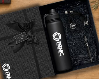 Könnte beinhalten: Eine schwarze Geschenkbox mit einer mattschwarzen Wasserflasche mit FBMC-Logo, einem schwarzen Kugelschreiber und einem schwarzen Multi-Port-Ladekabel. Der Deckel ist mit einer schwarzen Schleife und einem Anhänger mit dem Text Just For You versehen.