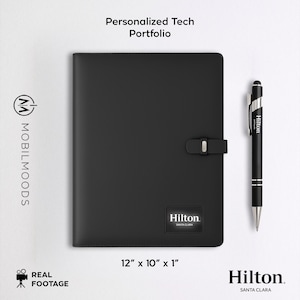 Può includere: Portfolio tecnologico personalizzato nero con penna. Il portfolio misura 30,5 cm x 25,4 cm x 2,5 cm, con chiusura a fibbia e il logo Hilton Santa Clara. Una penna nera con il logo Hilton è posizionata accanto al portfolio. Il testo "Personalized Tech Portfolio" è in alto.
